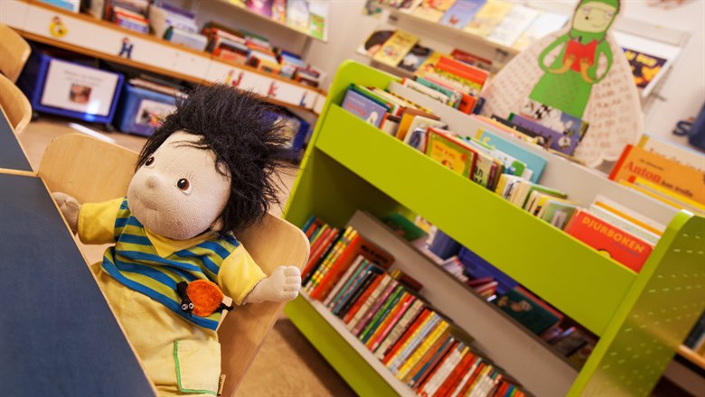En docka sitter vid ett bord. Grön hylla med barnböcker och Bokmalen i pappfigur i bakgrunden.