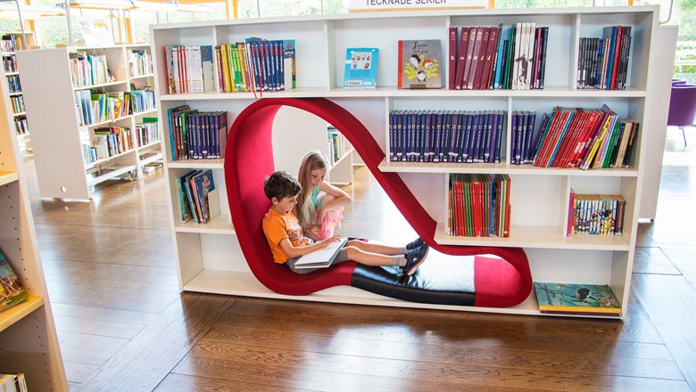Två barn sitter i en röd formation i en bokhylla på biblioteket och läser