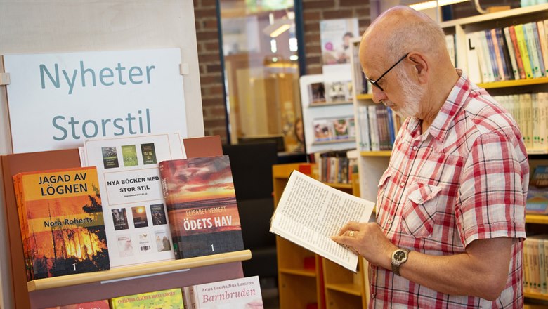 En äldre man står vid ett bokställ och läser ur en storstilt bok.  