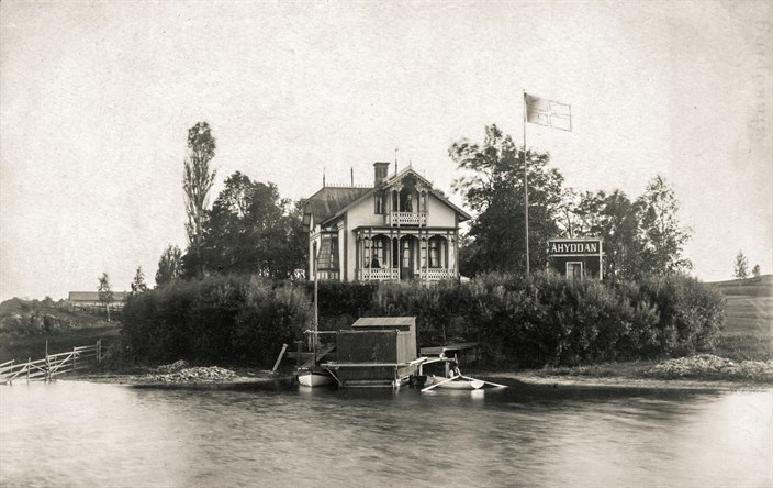 Svartvitt foto på en vacker träbyggnad som ligger intill vattnet. Bredvid huset syns en mindre byggnad som har texten Ålhyddan på fasaden.