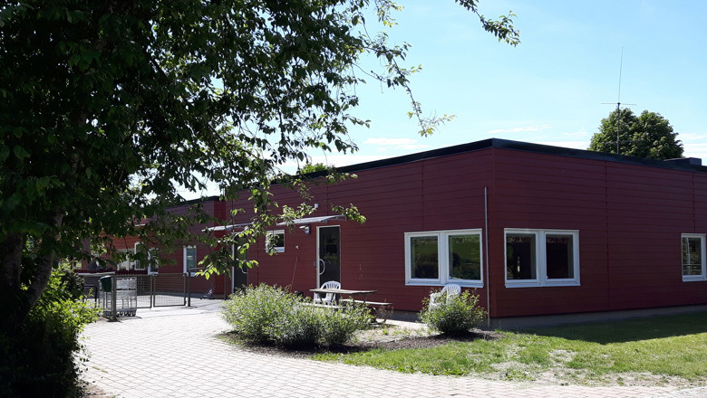 En röd förskolebyggnad inbäddad i gröna buskar och gräsytor, en stengång fram till huset.