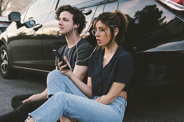 Två ungdomar sitter lutandes mot en bil och lyssnar på musik genom varsin hörlur.