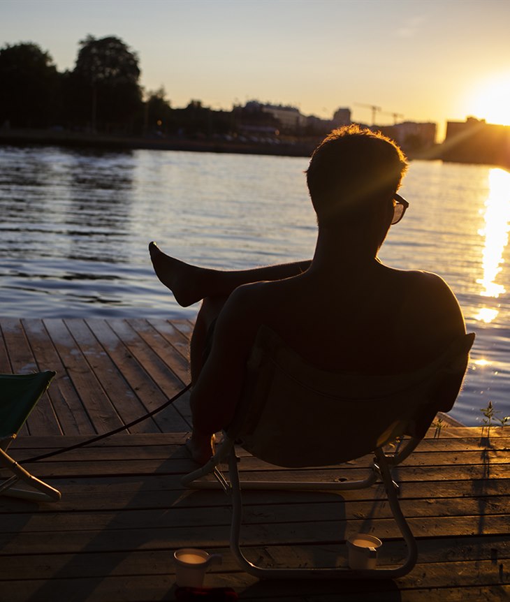 En kille sitter på en solstol på en brygga vid Stångån och njuter av solnedgången