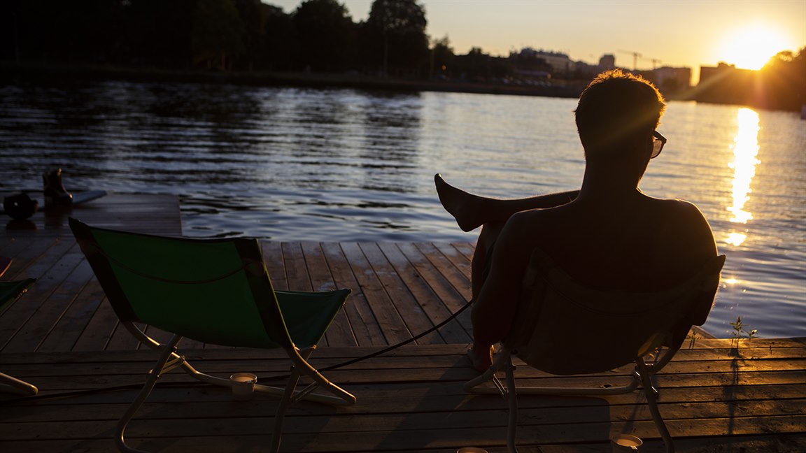 En kille sitter på en solstol på en brygga vid Stångån och njuter av solnedgången